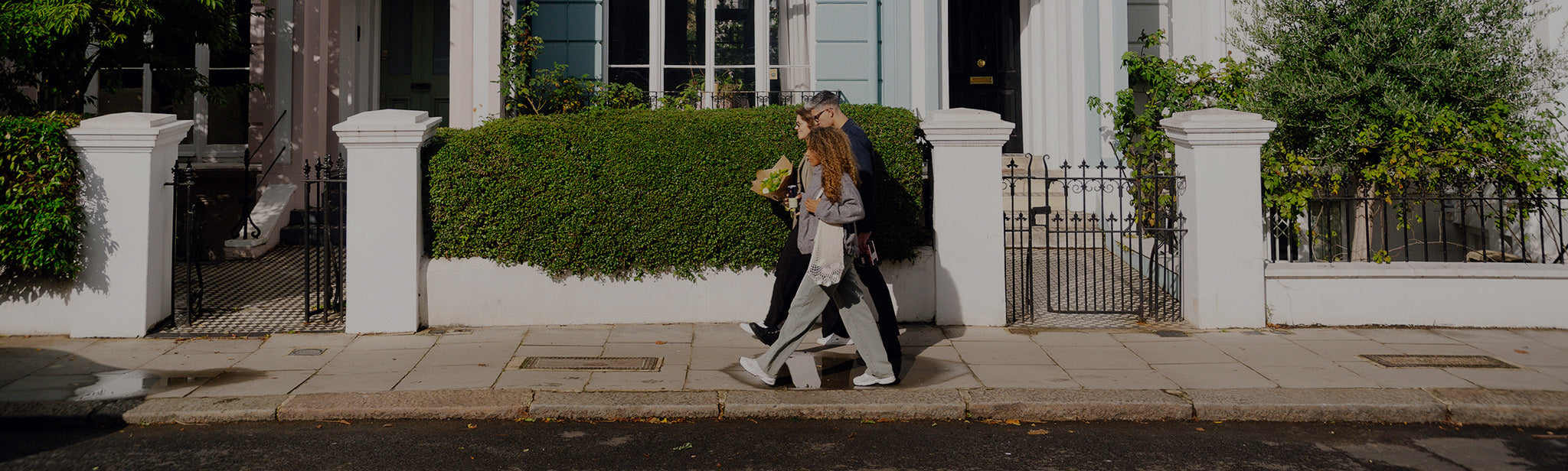 Three people walking on a sidewalk past white houses with green hedges on a sunny day.