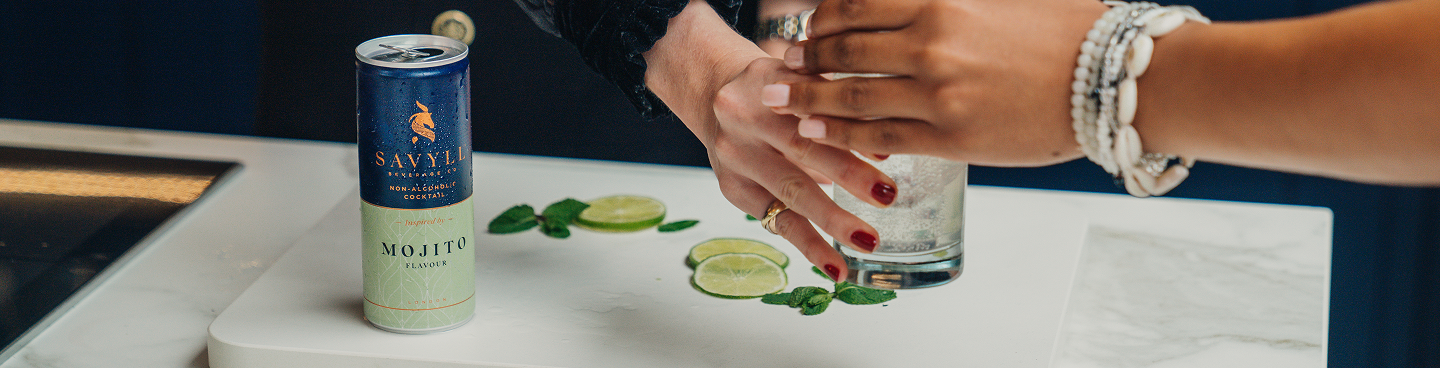 Can of Savvy non-alcoholic mojito cocktail next to hands holding a glass with lime slices and mint on a white surface.