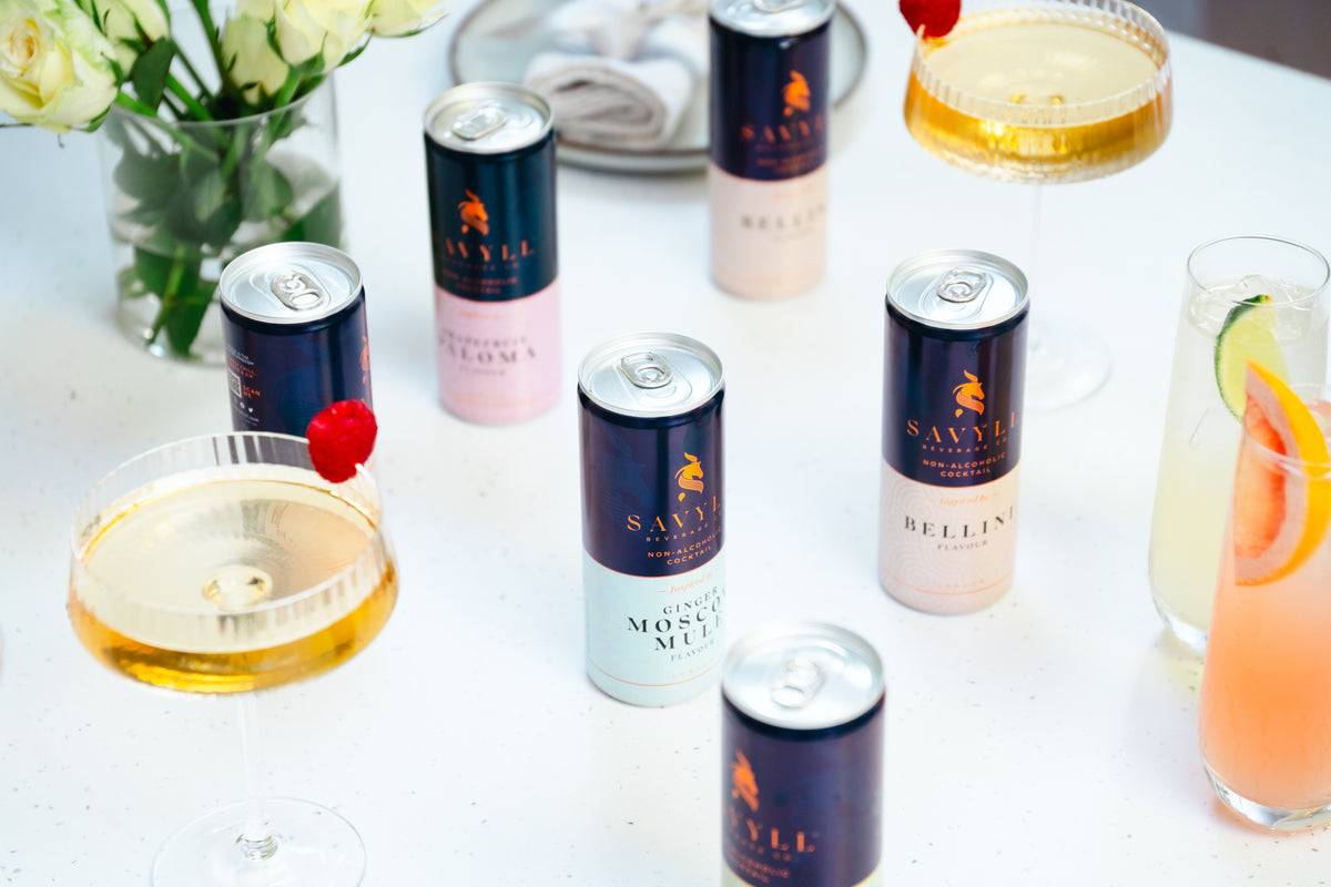 Cans of Savyll non-alcoholic cocktails in Bellini and Moscow Mule flavors on a white table with garnished drinks and flowers.
