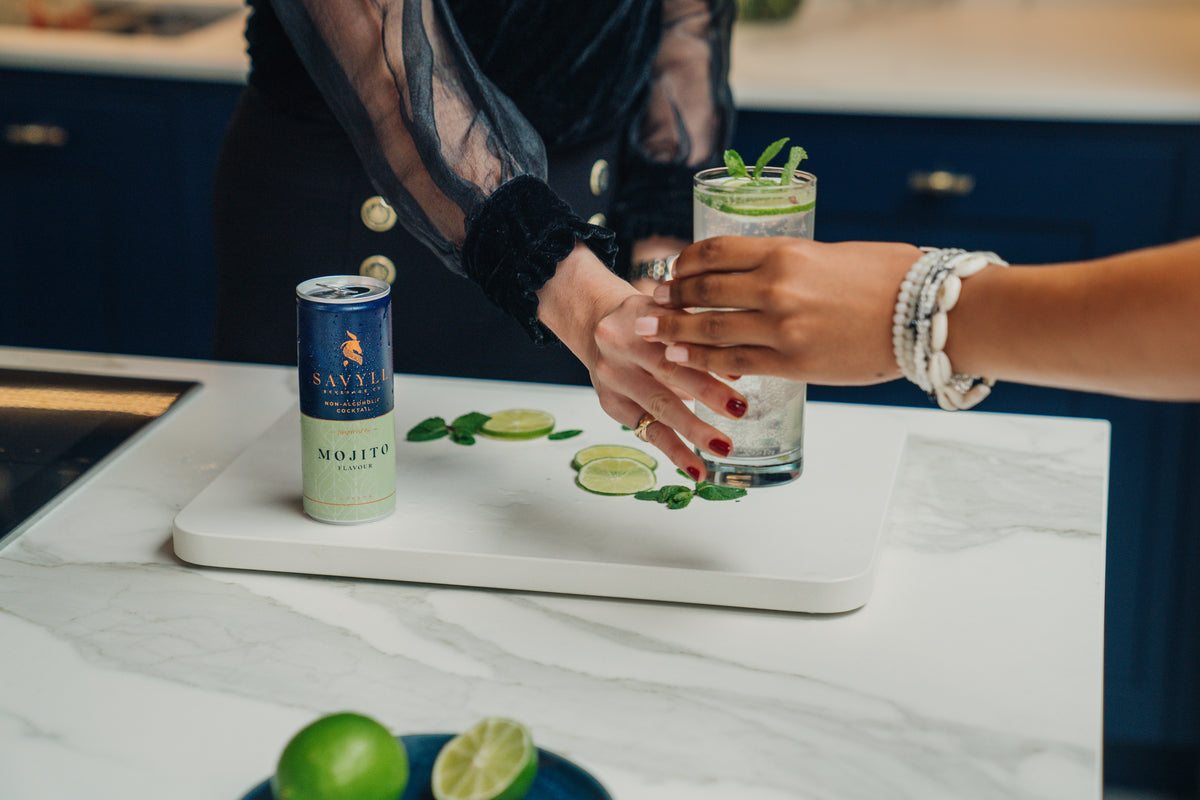 Two hands exchanging a mojito-flavored non-alcoholic cocktail in a glass with lime and mint on a kitchen counter.