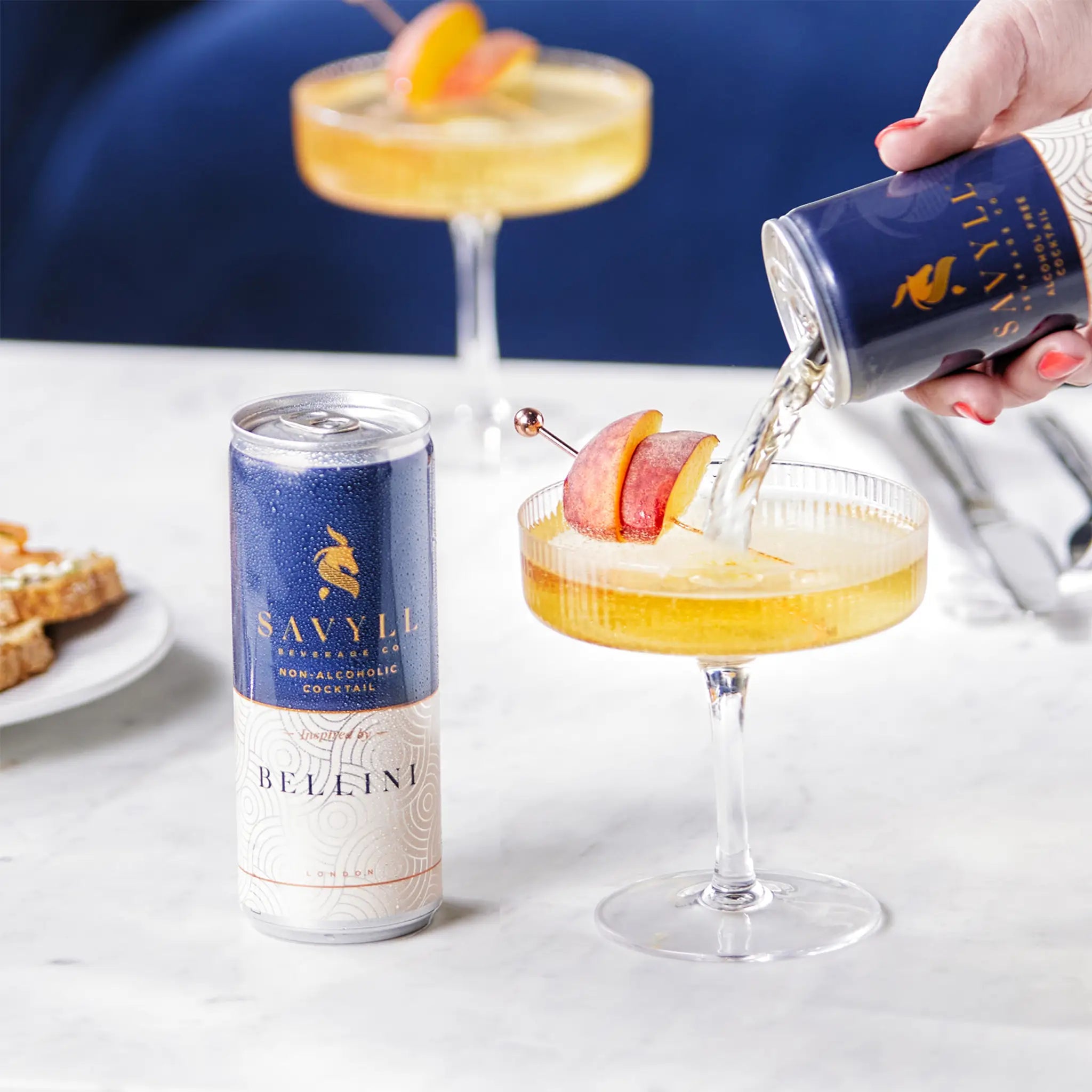 Hand pouring Savyll alcohol-free Bellini cocktail from can into glass garnished with peach slices on marble table.