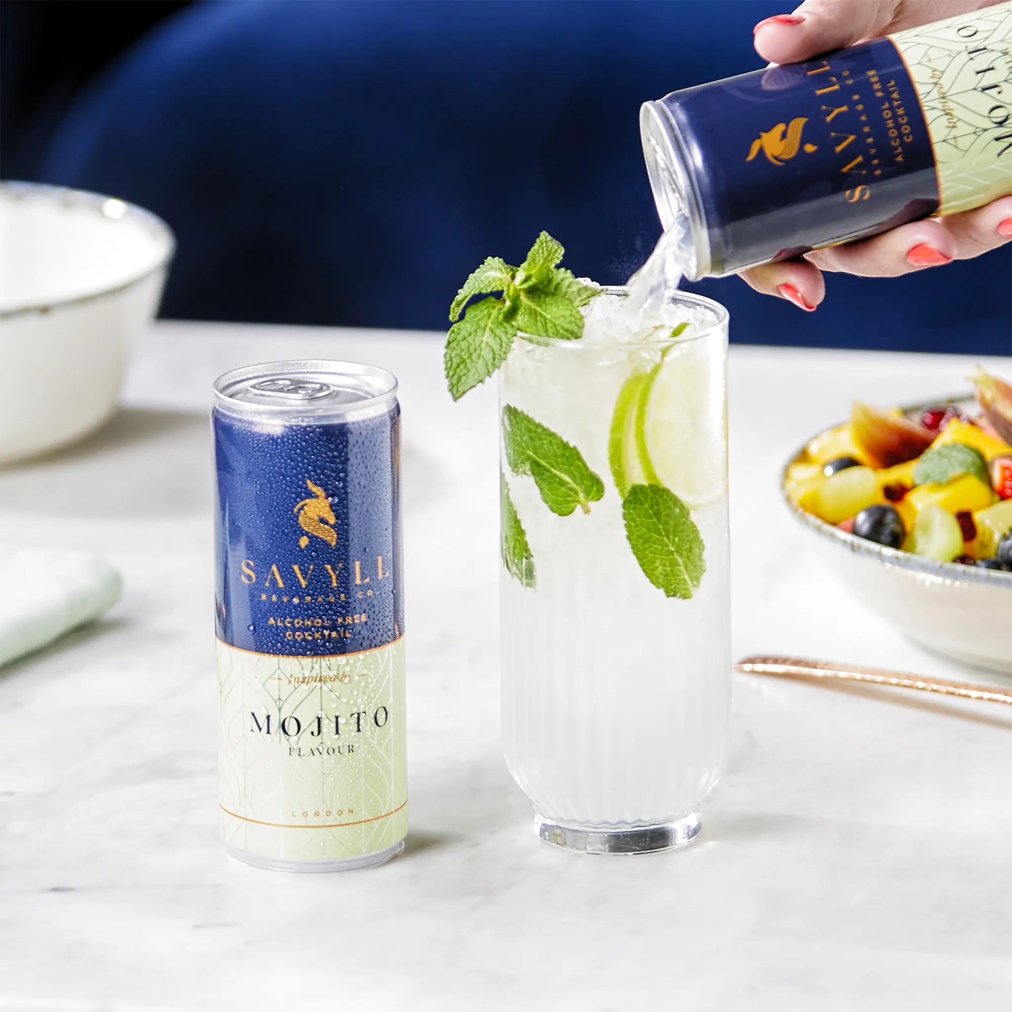 Can of Savyll alcohol-free mojito cocktail next to a glass with ice, lime, and mint being poured from another can.