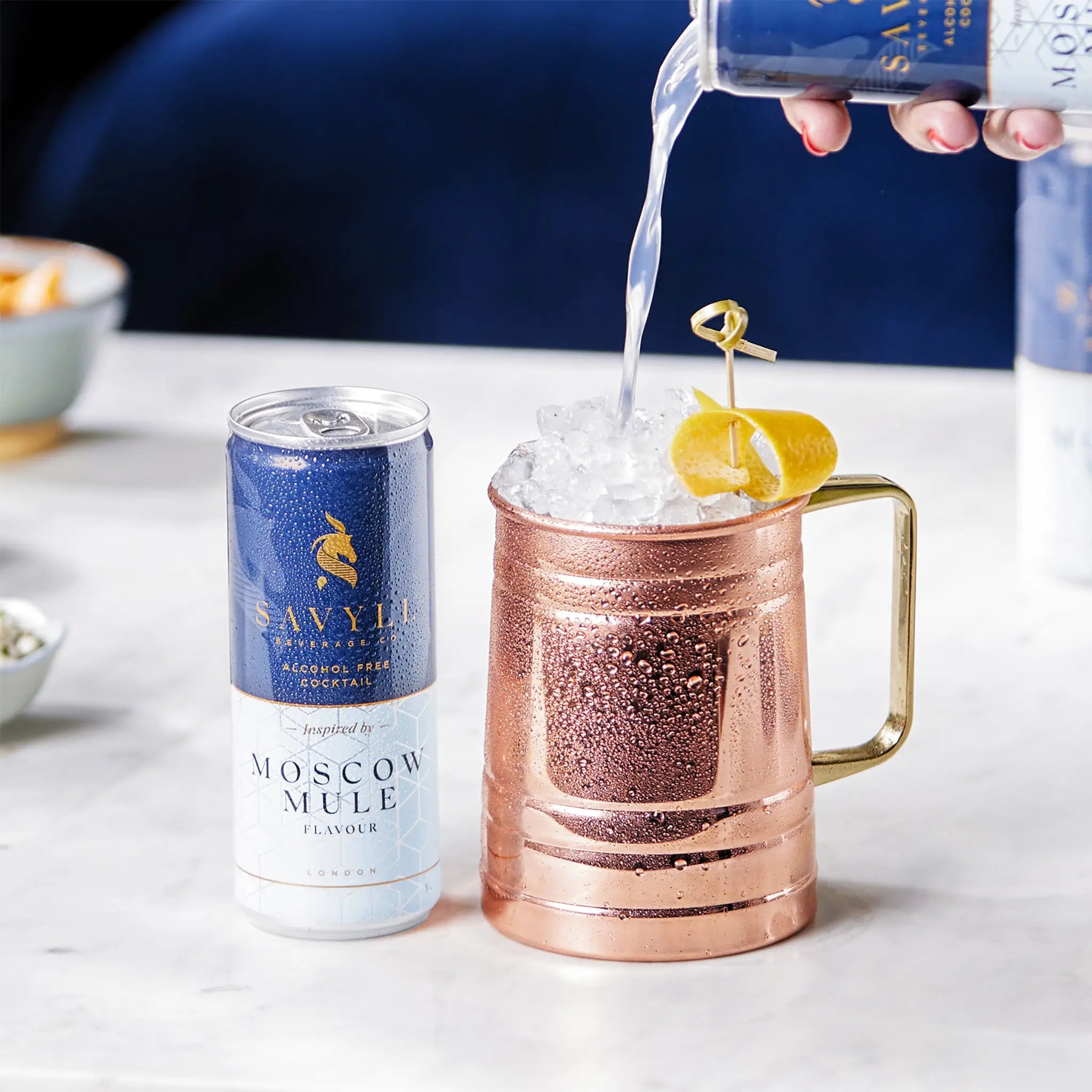 Can of Savyll alcohol-free Moscow Mule next to a copper mug filled with ice and lemon peel, drink being poured in.