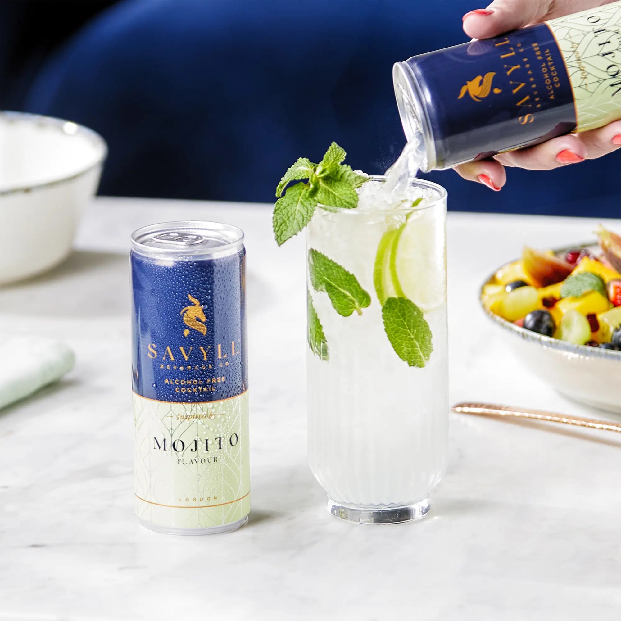 Hand pouring Savyll alcohol-free mojito from can into glass with lime slices and mint on marble table.